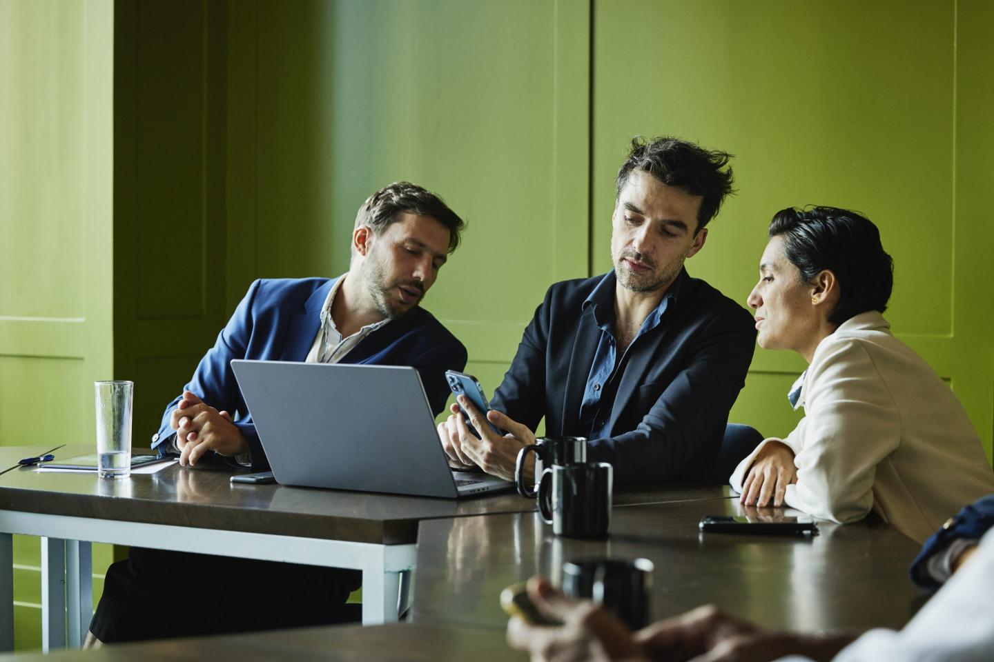 Three people sit behind a desk and look at the phone screen of the person in the middle.