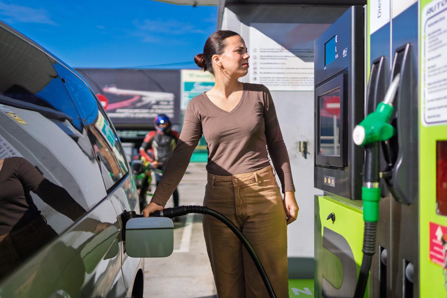 Woman refuels her car at a gas station on a sunny day, pondering rising gas prices and environmental impact. Transportation, energy, and the environment intersect in daily life