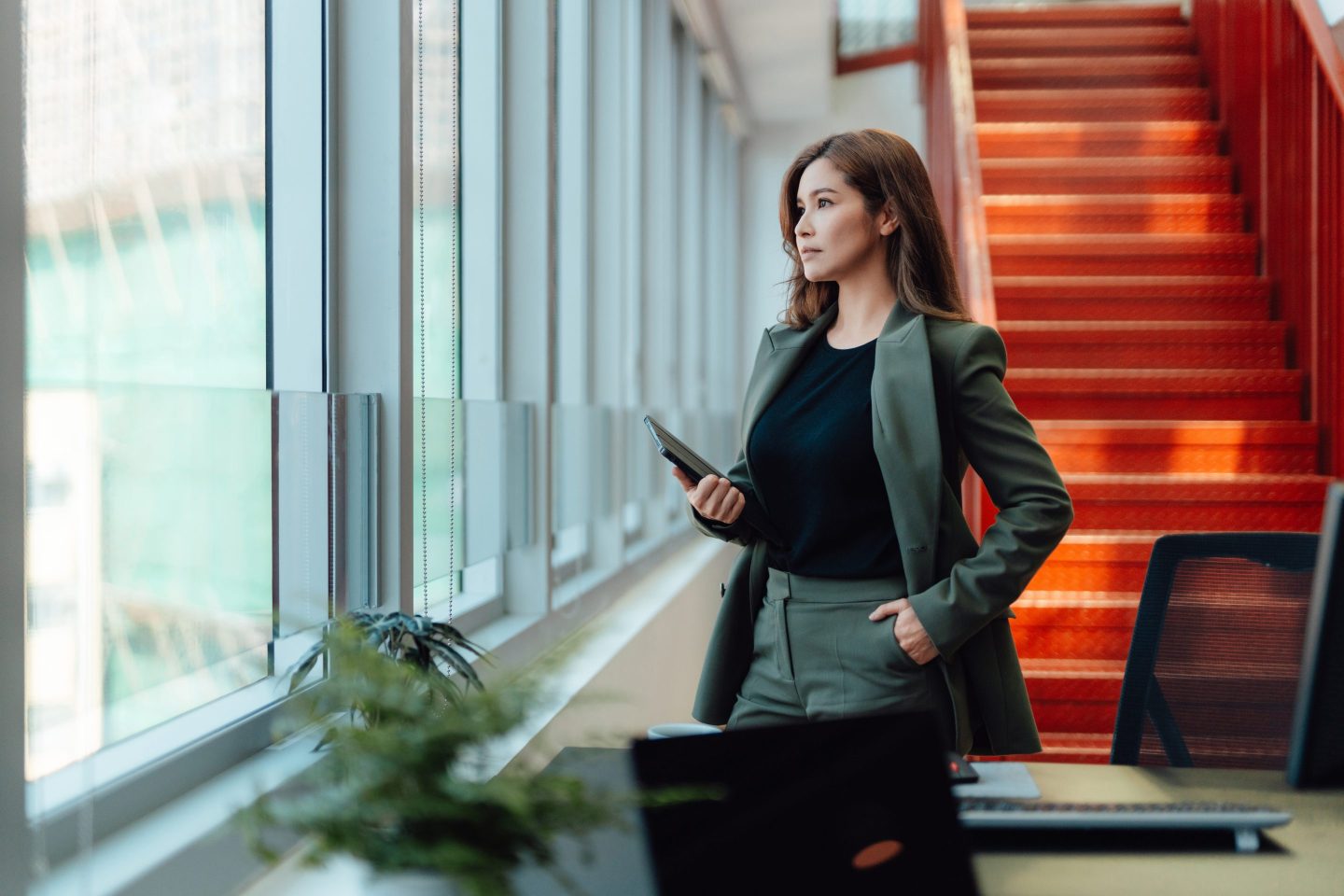 Thoughtful businesswoman holding a digital tablet, looking through window, setting goals and thinking about career progression, standing by the window of a bright and modern office. Women's empowerment and career achievement.