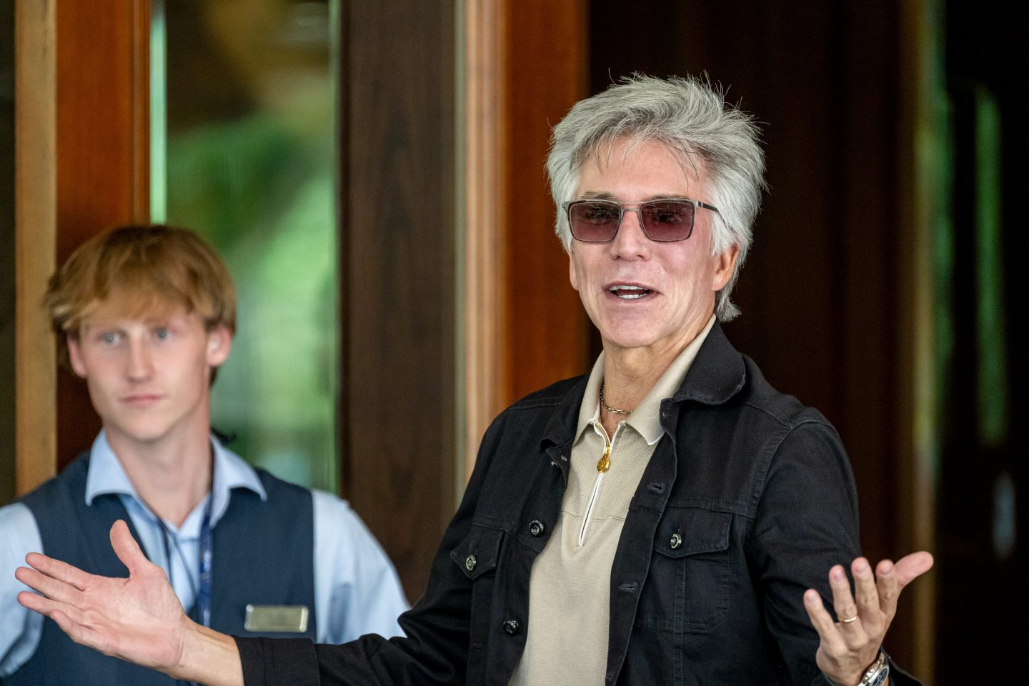 ServiceNow CEO Bill McDermott, wearing a casual jacket over a tope shirt and sporting sunglasses, raises his arms in a shrug-like gesture.