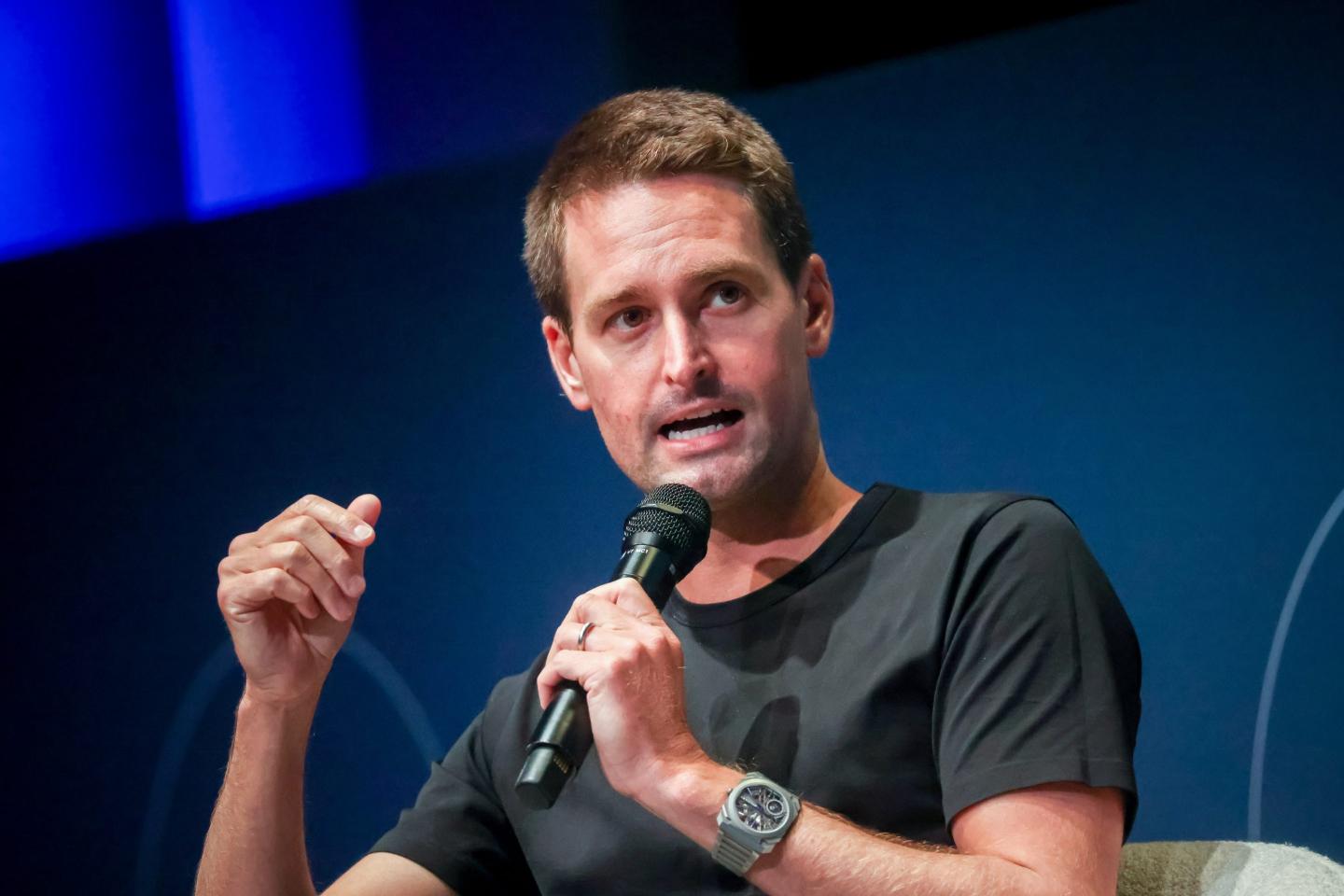 Evan Spiegel wears a black t-shirt and speaks into a microphone while on stage.