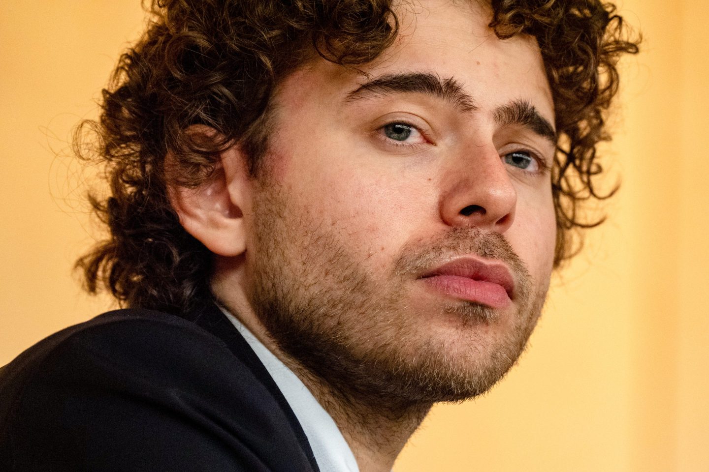 Shayne Coplan, in front of a yellow background, looks in front of him.
