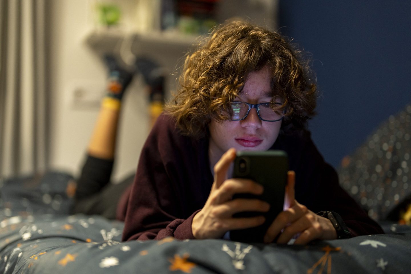 A teen boy lays on a bed looking at his phone.