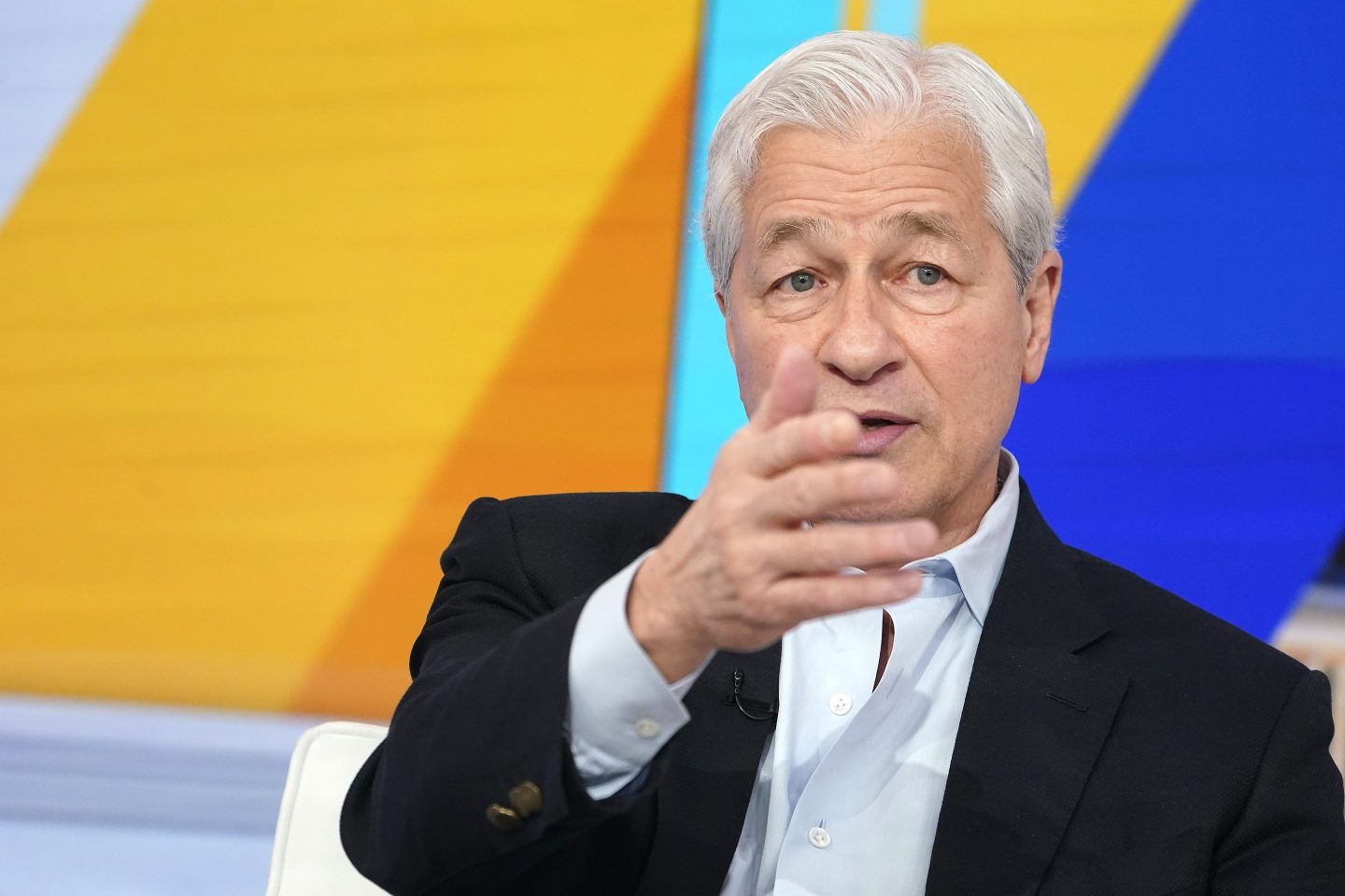 Jamie Dimon, in front of a colorful background, gestures with one hand outstretched, covering part of his face.