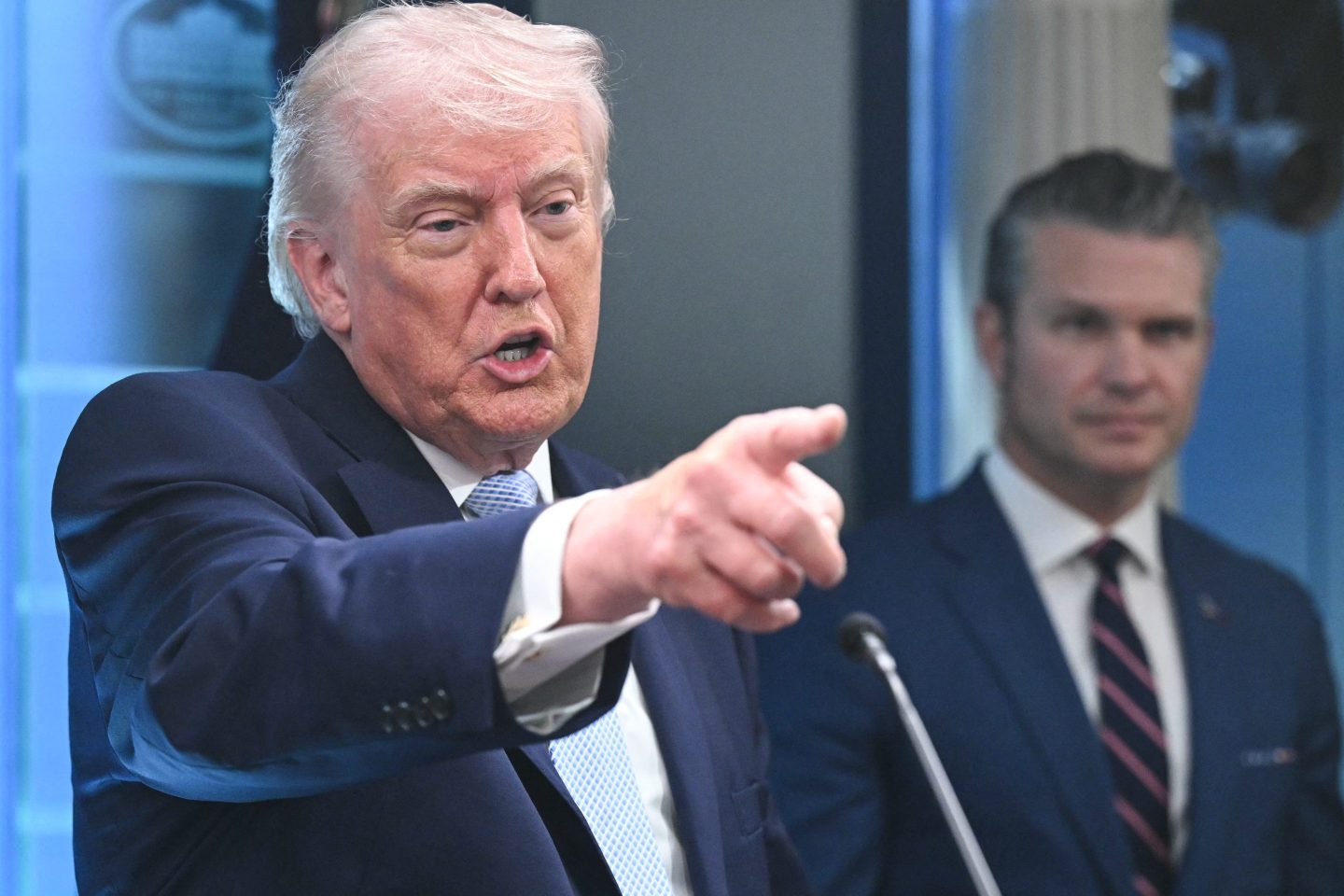 Donald Trump points and speaks as Pete Hegseth looks on in the background.