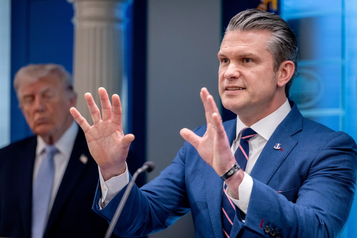 Pete Hegseth speaks with both hands in the air as Donald Trump looks on in the background.
