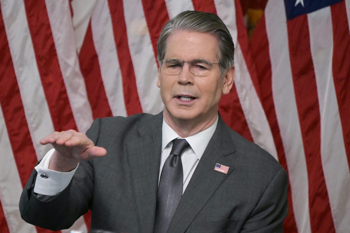 Scott Bessent, standing in front on an American flag, puts one hand up as he talks.