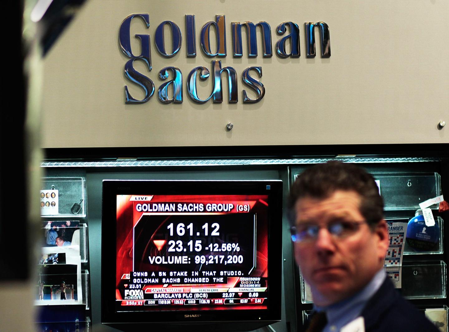 A television screen showing Goldman Sachs' stock price plays in front of a Goldman Sachs sign