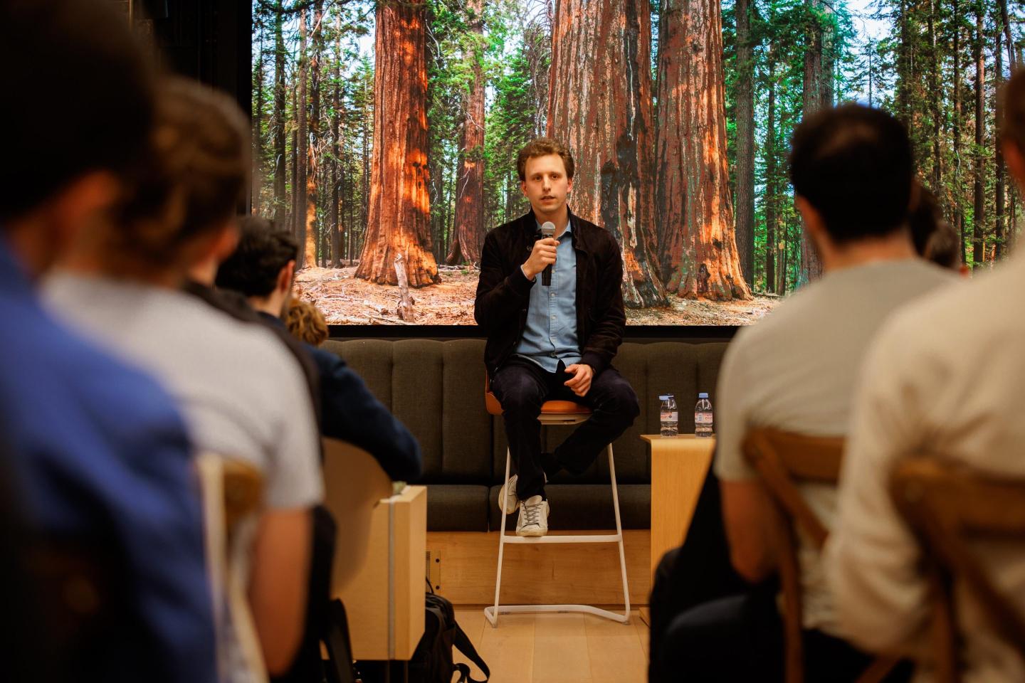 Sequoia partner Julien Bek sitting on a stool and holding a microphone while speaking to an audience. Behind him is a stage that looks like a forest.