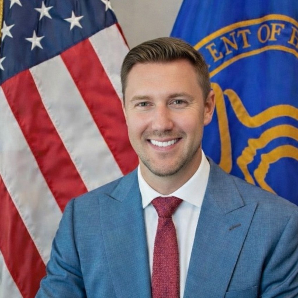 man in gray suit before American and HHS flags.