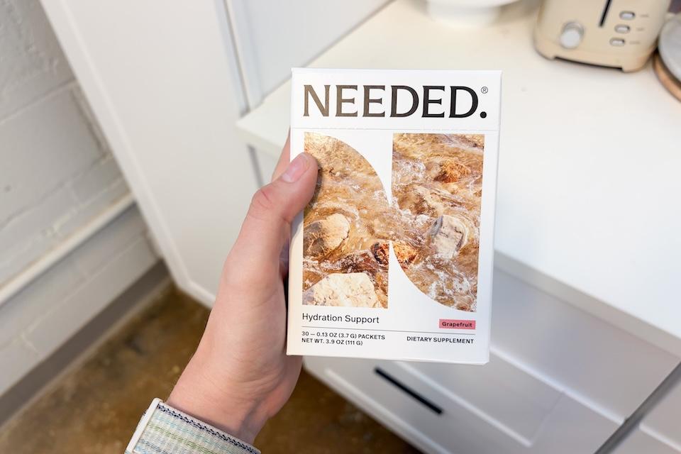 A hand holding a box of Needed Hydration Support. The packaing is a white box with an image of running water in a stream on the front.