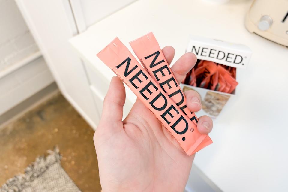 A closeup of a hand holding two individual packets of Needed Hydration Support. The packets are a salmon-pink color.
