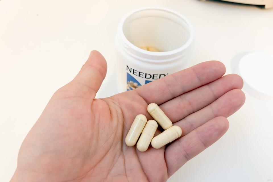 An open palm holding four Needed Women's Multivitamin capsules. On a countertop below, you can see an open bottle containing more of the capsules.