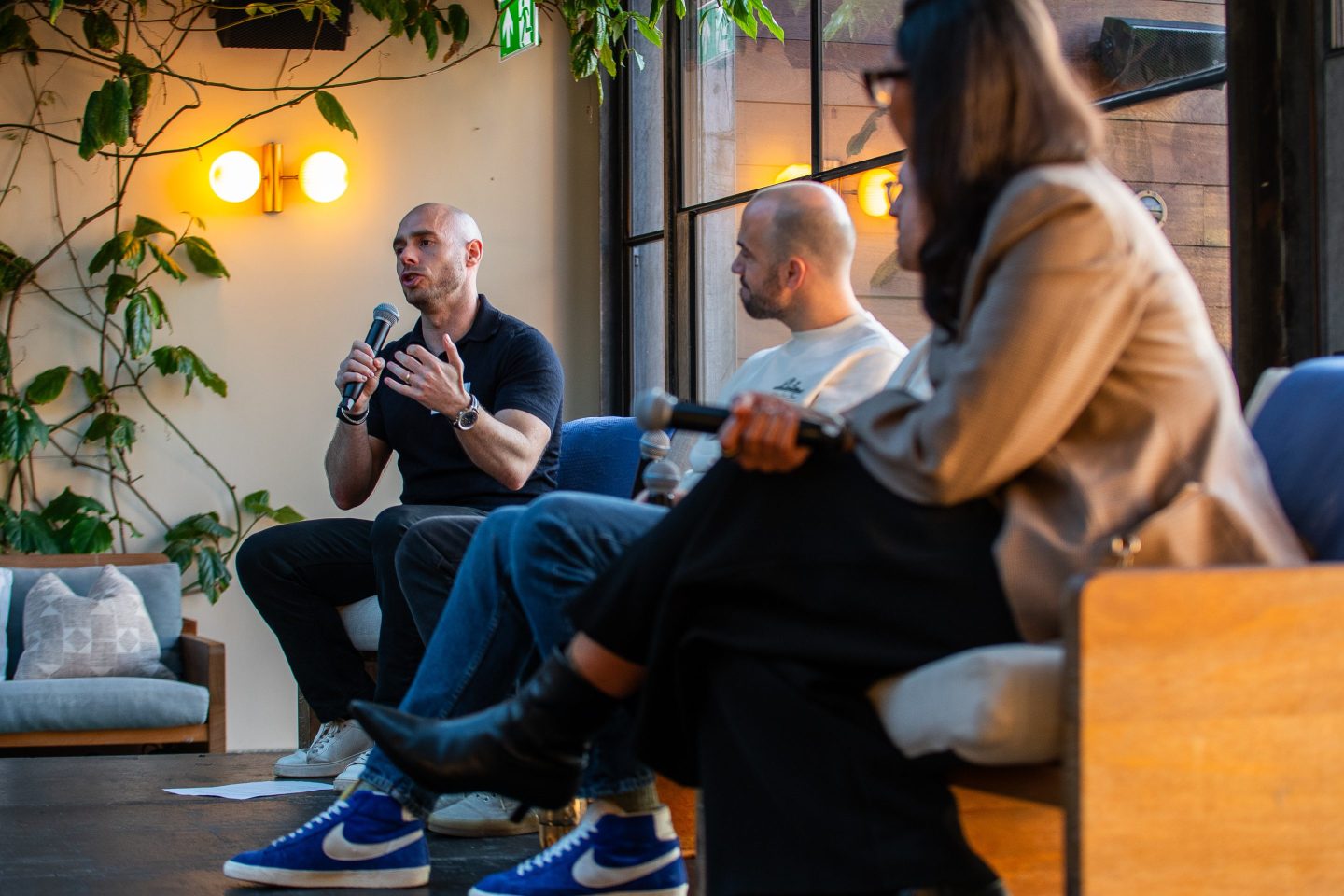Dex founder and CEO Paddy Lambros (left) sits on a stool on a stage with two other panelists while holding a microphone and gesturing.