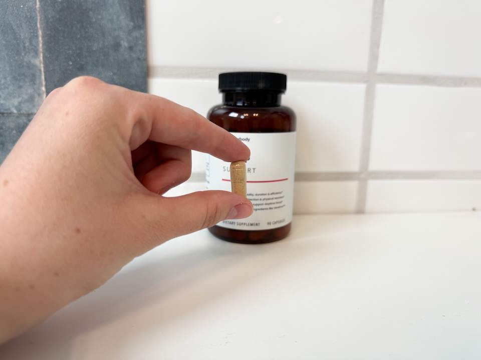 A hand holding an Innerbody Labs Sleep Support capsule between the index finger and thumb.