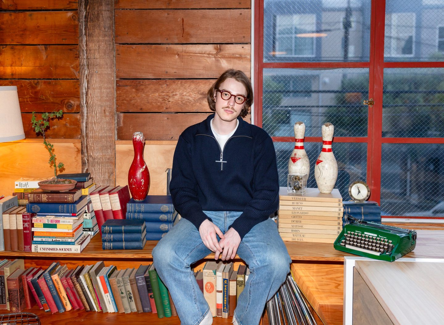 Paul Scherer poses casually while sitting on a bookshelf