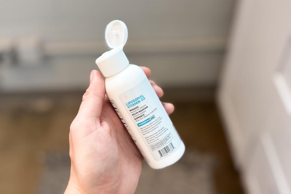 A hand holding a bottle of 1st Phorm Liposomal Vitamin D3. The cap is flipped open.