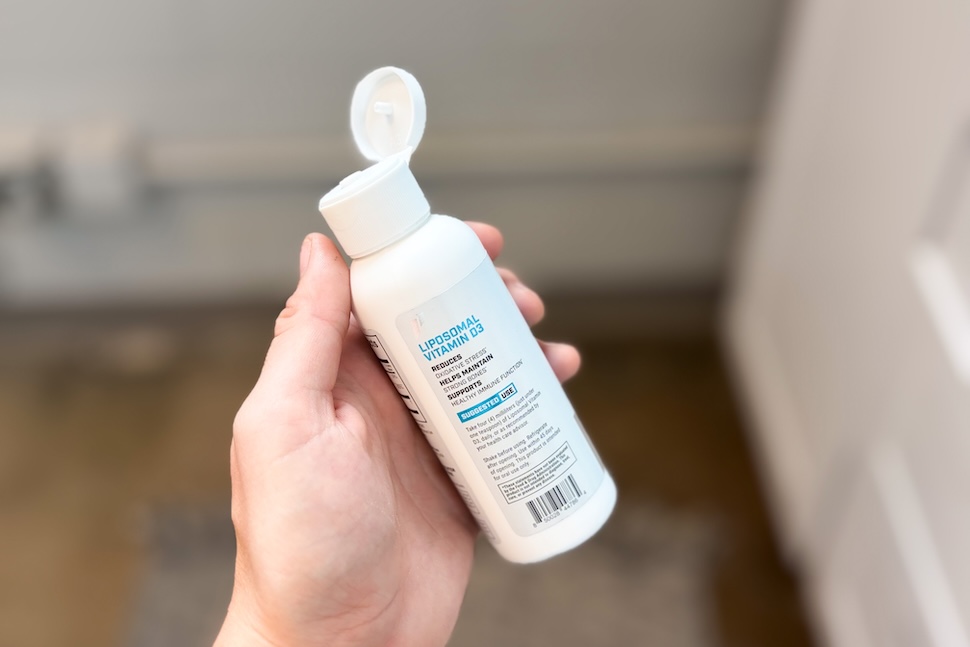 A hand holding a bottle of 1st Phorm Liposomal Vitamin D3. The cap is flipped open.