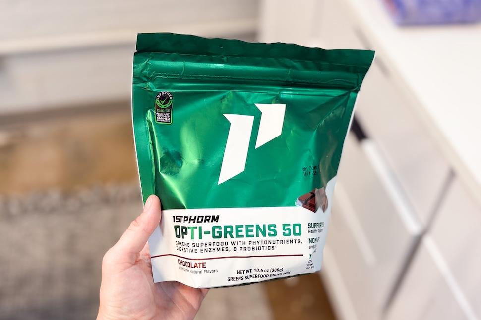 A hand holding a pouch of 1st Phorm Opti Greens 50. The packaging is a shiny green and white.