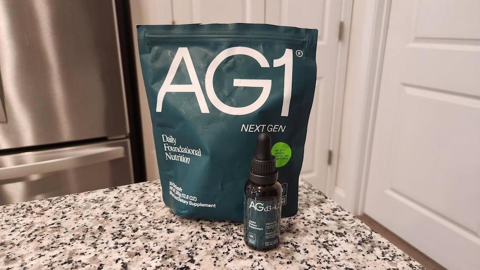 A bottle of AG1 Vitamin D3+K2 sitting in front of a pouch of AG1 on a marble countertop