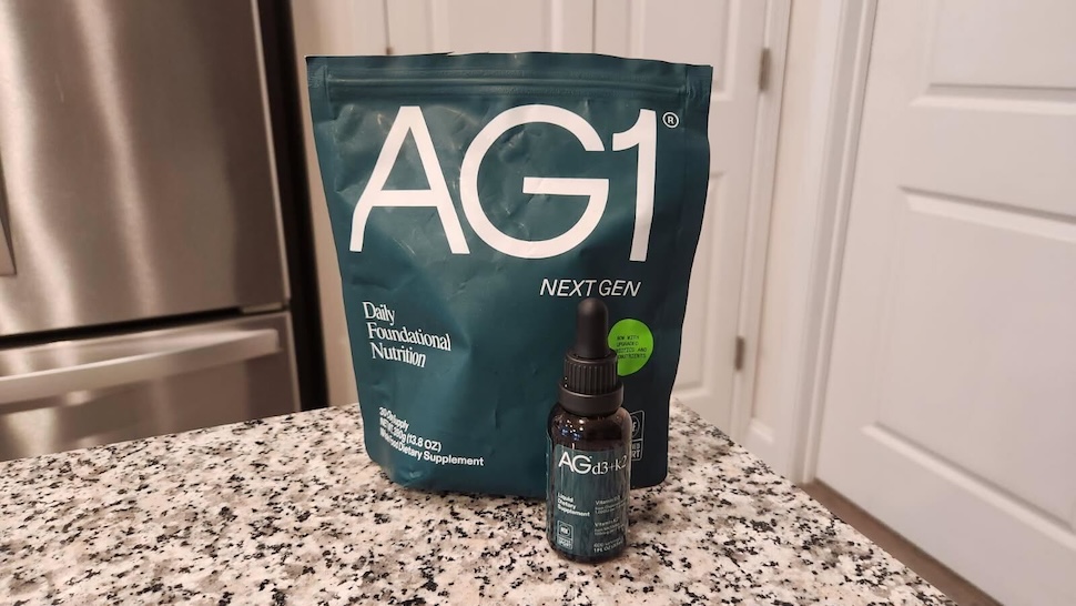 A bottle of AG1 Vitamin D3+K2 sitting in front of a pouch of AG1 on a marble countertop