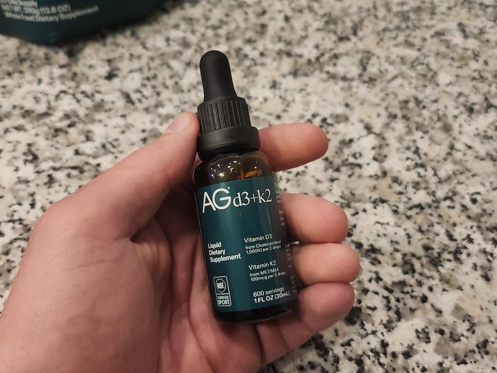 A hand holding a bottle of AG1 D3+K2