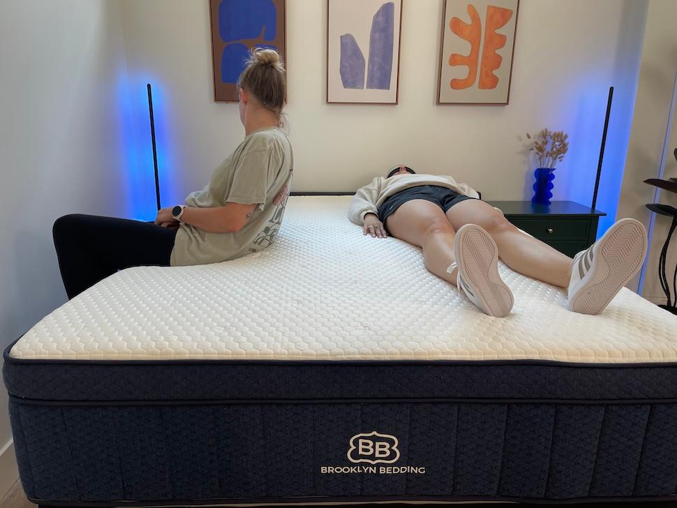A woman sitting on the edge of the Brooklyn Bedding Aurora Luxe mattress. Another woman lies next to her, wearing a sleeping mask.