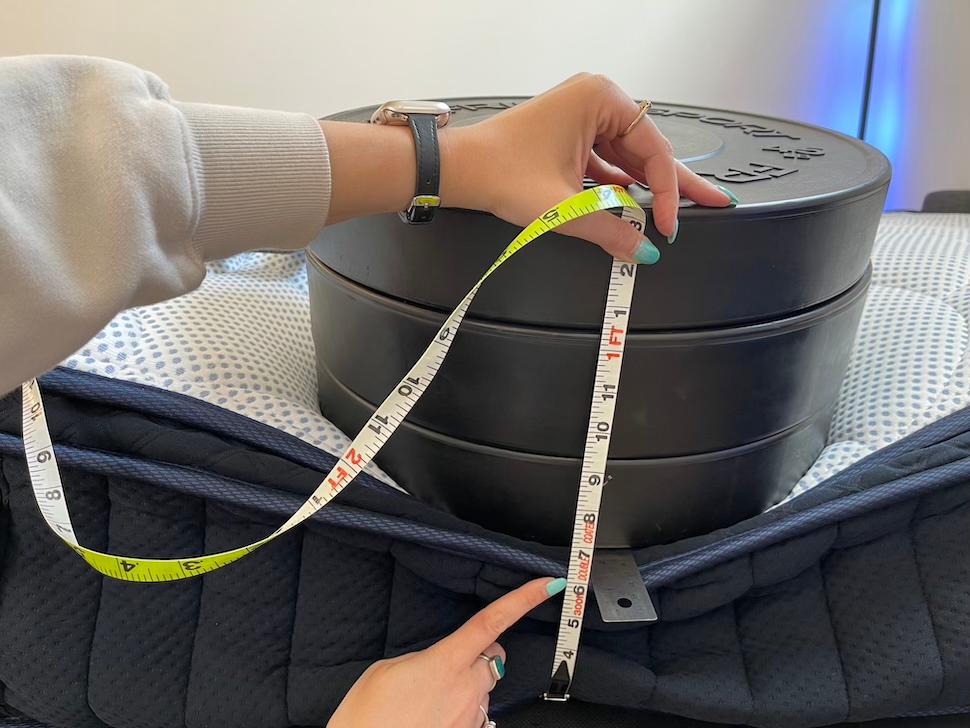 A stack of weight plates resting on the edge of a DreamCloud Premier Memory Foam mattress. A woman is kneeling and measuring the sinkage caused by the weights.