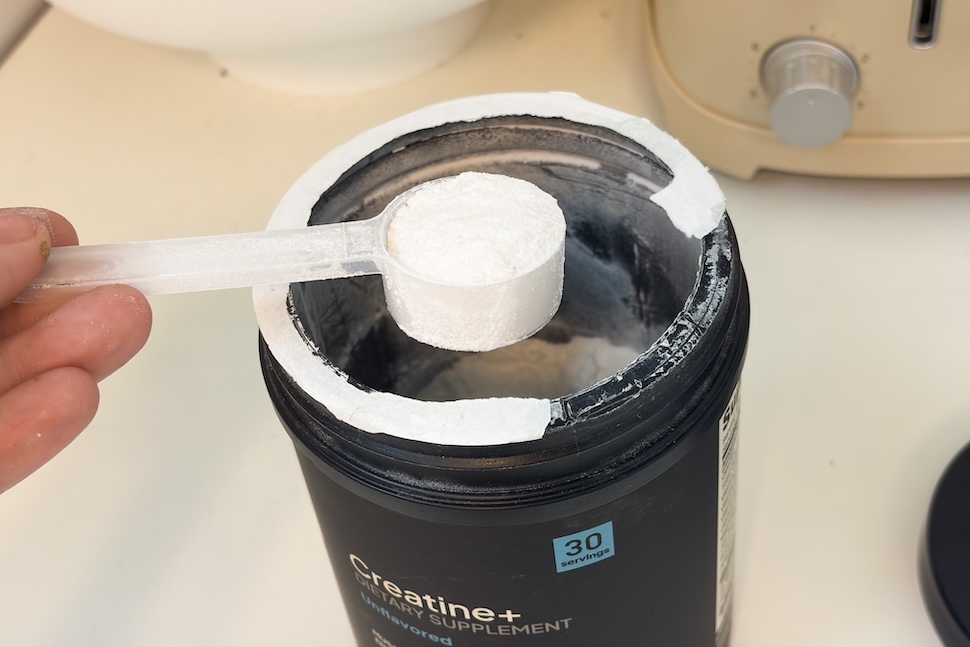 An open canister of Factor Form Creatine + Electrolytes. A hand is using a plastic scoop to scoop out some of the fine white powder.