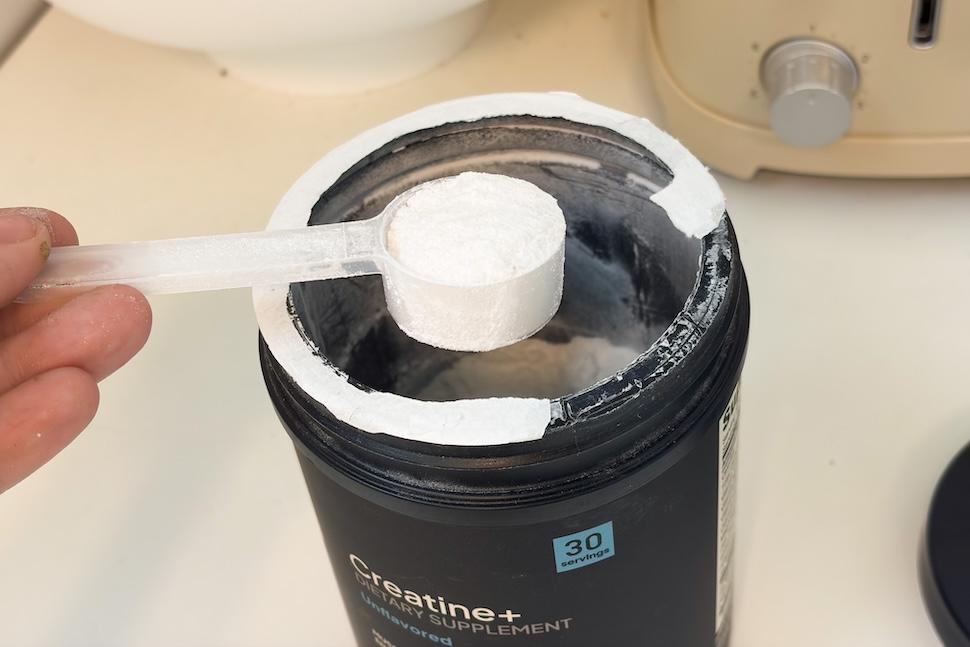 An open canister of Factor Form Creatine + Electrolytes. A hand is using a plastic scoop to scoop out some of the fine white powder.