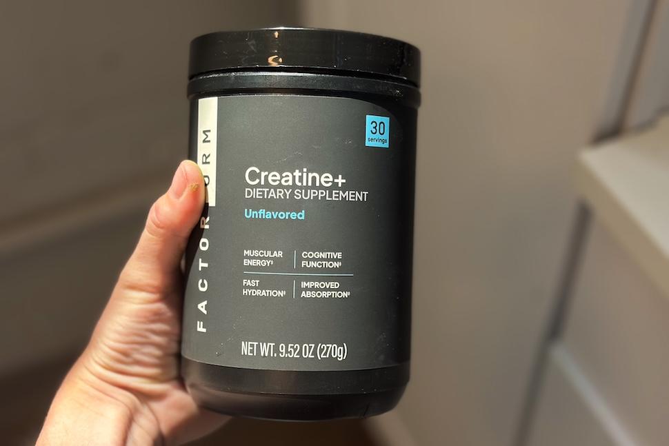 A hand holding up a black canister of Factor Form Creatine + Electrolytes