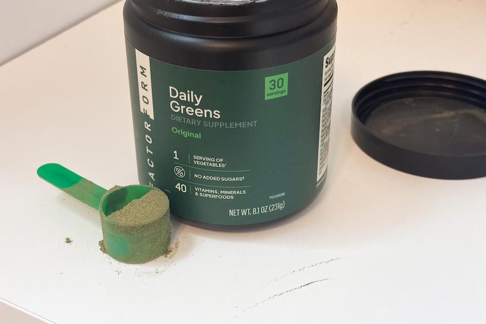 A canister of Factor Form Daily Greens sitting on a white countertop. Next to it is a green scooper filled to the top with greens powder.