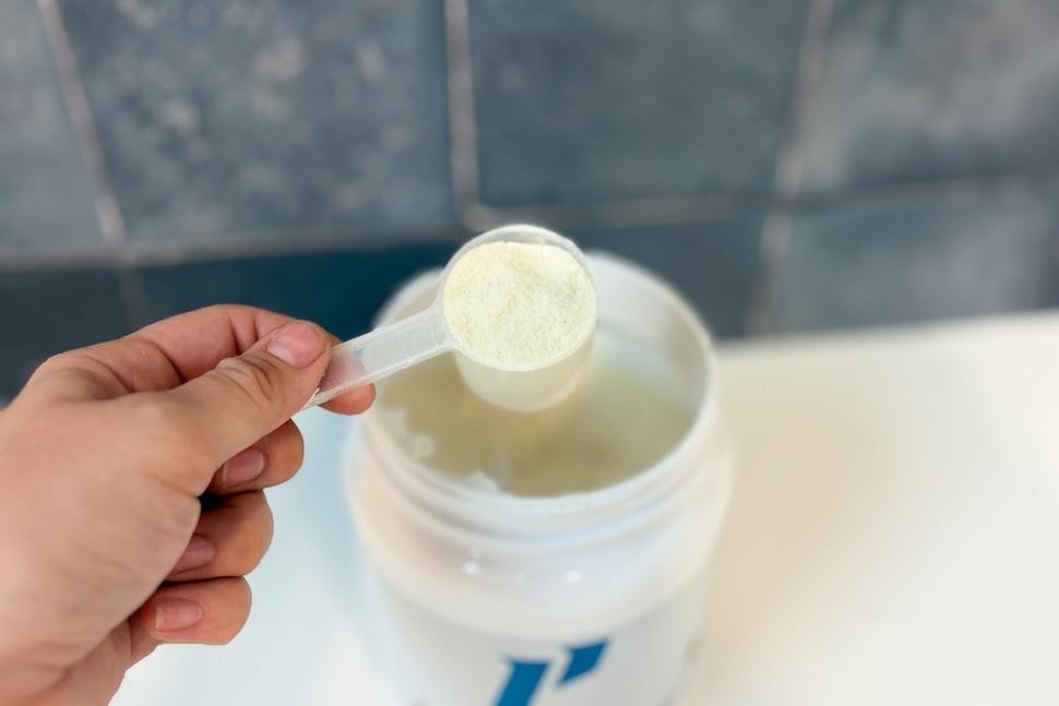 An open canister of 1st Phorm Collagen sits on a white countertop. A hand has used a scooper to take out one scoop of the off-white powder.