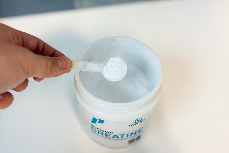 An open canister of 1st Phorm Micronized Creatine Monohydrate. A hand is reaching in with a plastic scoop and taking out one scoop of the fine white powder.