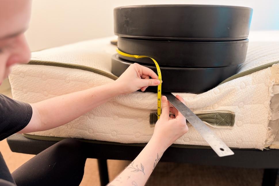A stack of weight plates on the edge of an Avocado Green mattress. A woman is crouching next to the weights and measuring their sinkage with a tape measure.