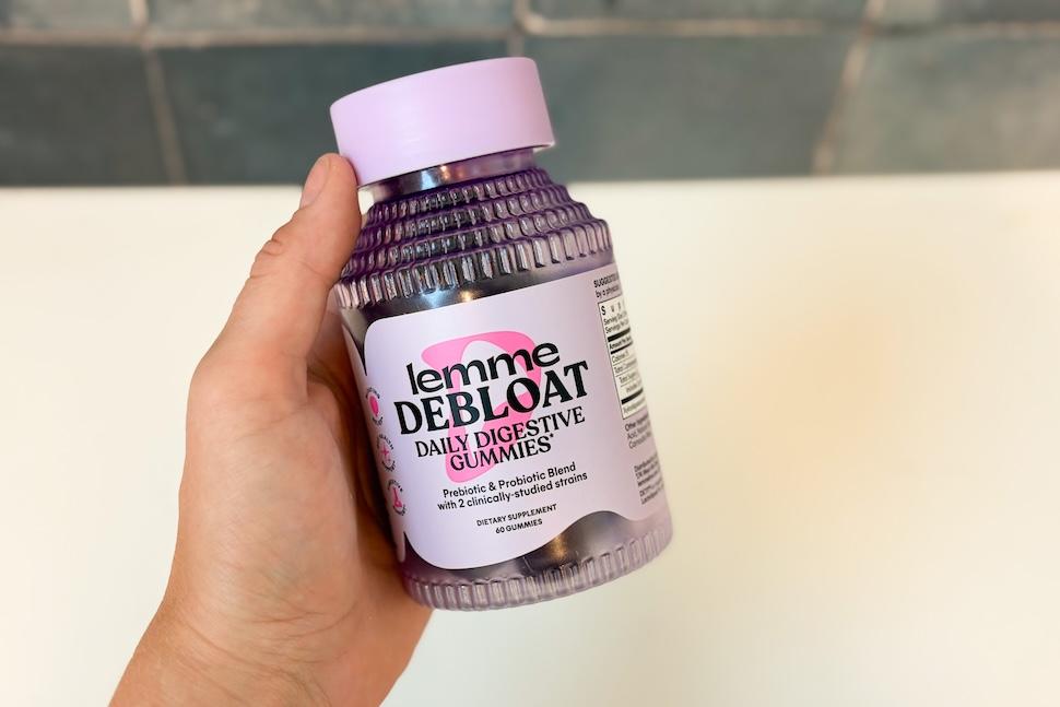 A hand holding a jar of Lemme Debloat Daily Digestive Gummies. The bottle is a transparent purple with a lilac cap.