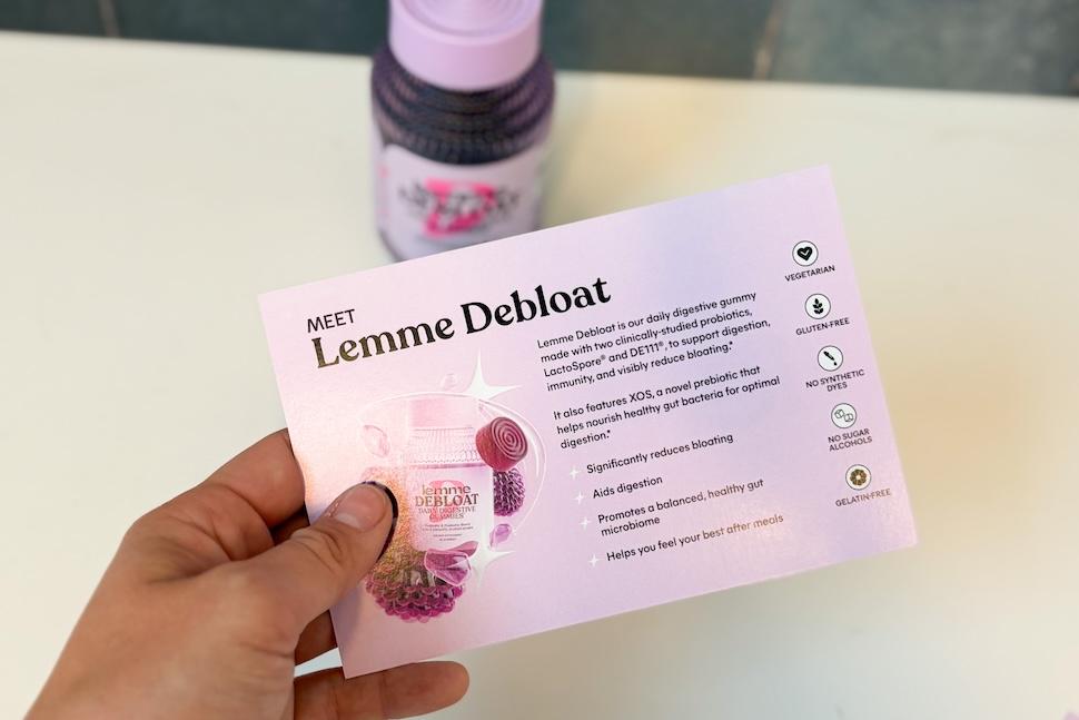 A closeup of a hand holding up a card displaying the benefits of Lemme Debloat gummies. On the countertop below is a purple bottle of the supplements.