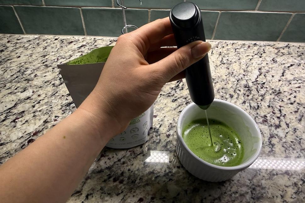 A hand using a mixer to stir a mug of Mantra Matcha Super Premium Ceremonial Matcha
