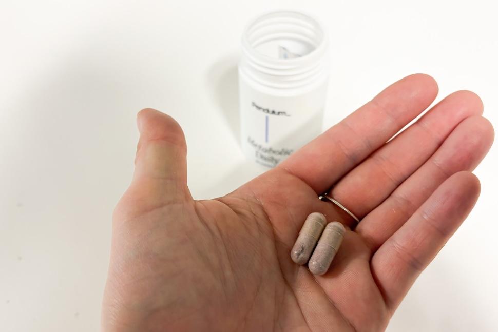 An open palm holding two Needed Pendulum Metabolic Daily capsules. On the countertop below, an open bottle of the supplements is visible.