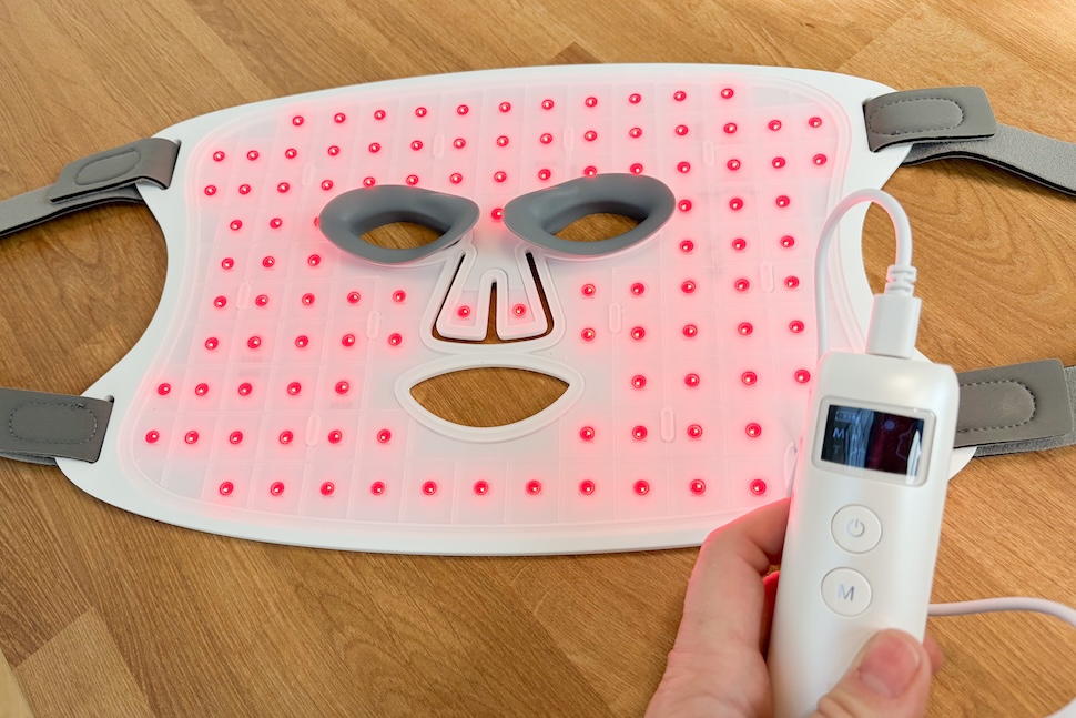 The Novaalab Glow Therapy Mask lying on a wooden table. A hand is holding a control connected to the mask.