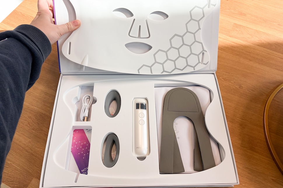 A closeup of a hand opening the box of the Novaalab Glow Therapy Mask. Inside, we see the mask, as well as its accessories, including a control and cord.