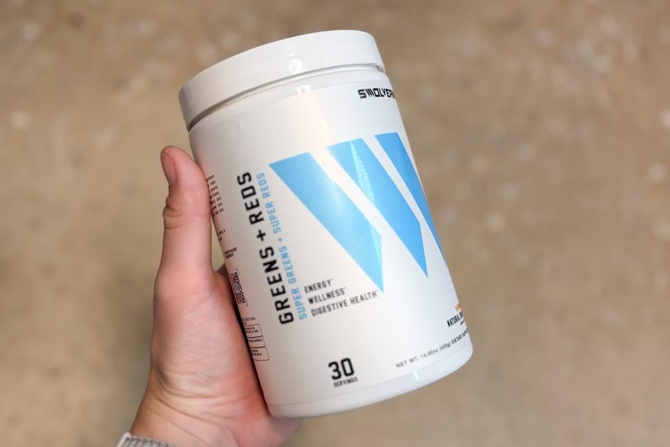 A hand holding a canister of Swolverine Greens + Reds Powder