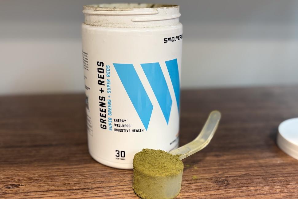 A canister of Swolverine Greens + Reds on a wooden countertop. A scooper packed to the top with the powder sits next to it.
