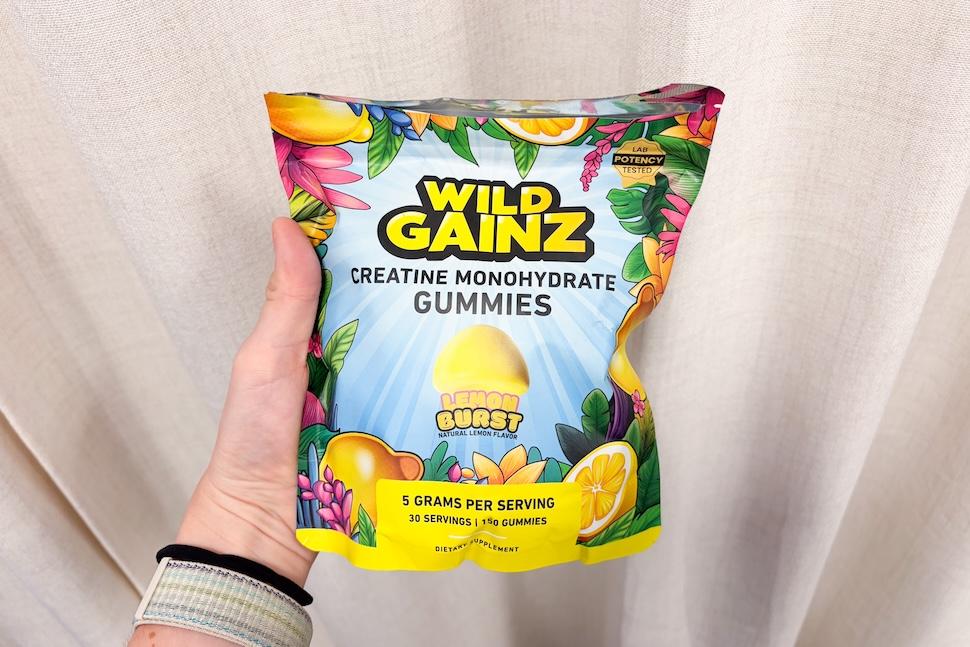 A hand holding up a bag of Wild Gainz Lemon Burst Gummies. The bag is colorful, with drawings of tropical flowers and fruit surrounding the letters.