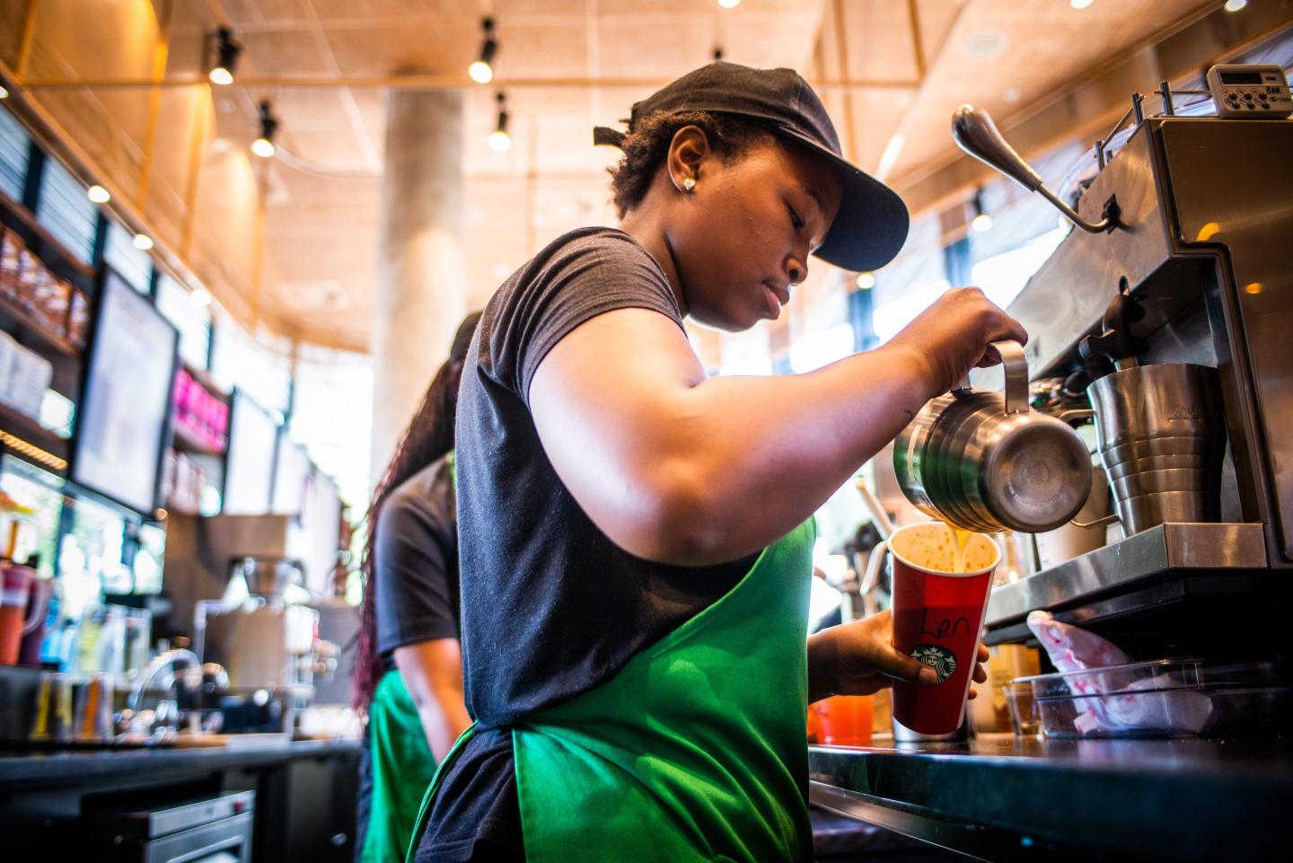 A barista wearing a green apron stands behind the bar and pours a drink into a cup