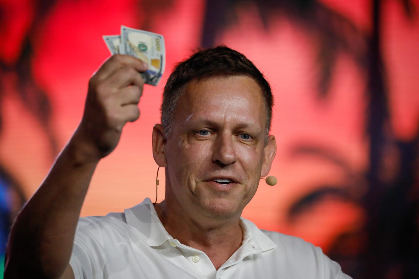 Peter Thiel, wearing a white shirt and in front of a red background, holds up a dollar bill.