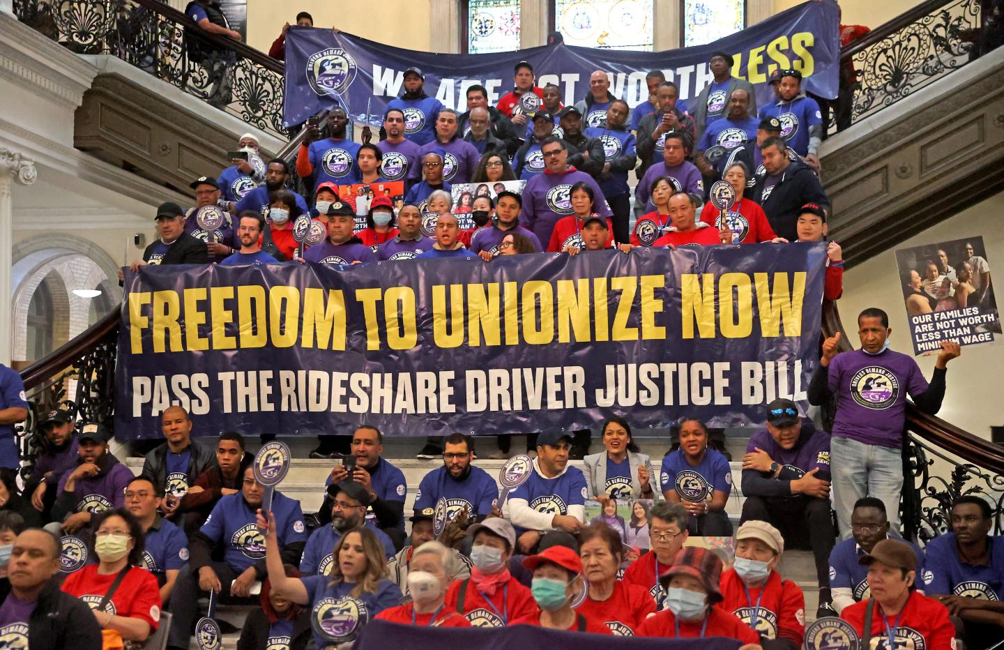 Dozens of people sit on a stairs and hold a large banner reading 'Freedom to unionize now'