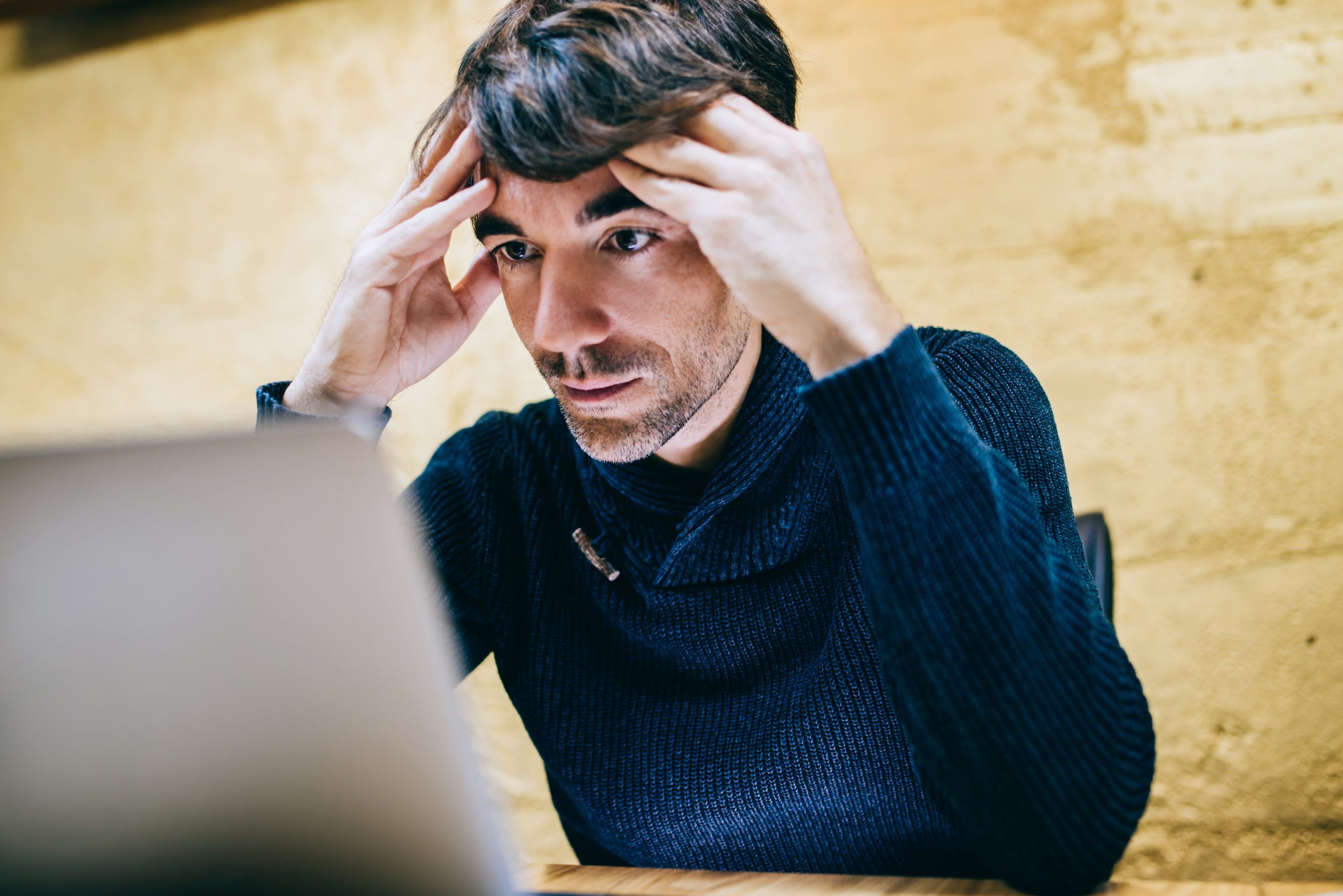 A man looks at this laptop and frowns, with both hands on the side of his head.