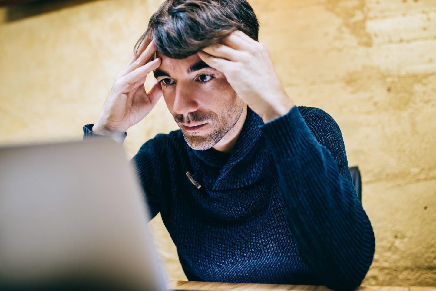 A man looks at this laptop and frowns, with both hands on the side of his head.