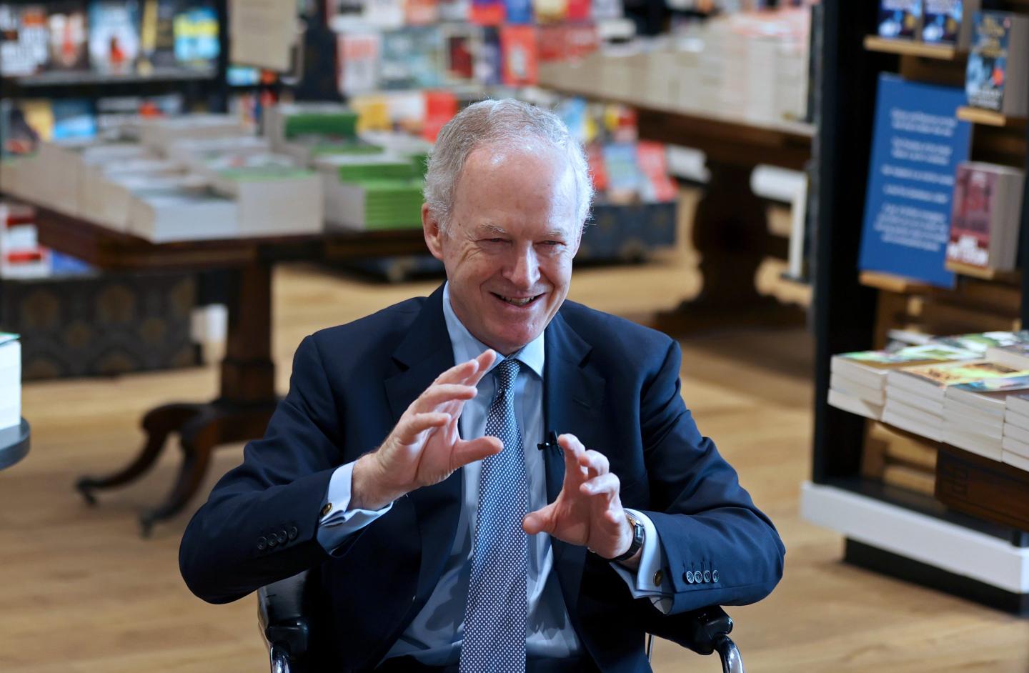 James Daunt sits in a booksop, gesturing with both hands and smiling.